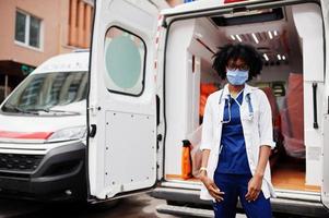 paramédica afro-americana em máscara médica protetora facial em frente ao carro da ambulância. foto