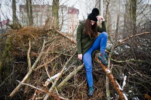 jovem usa moletom verde longo, jeans e chapéu preto em galhos do pinheiro em dia de inverno. foto