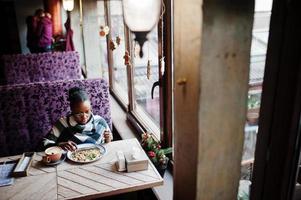 mulher africana em capa quadriculada e óculos sentado no café e comendo sobremesa. foto