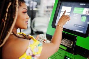 linda garota afro-americana de pequena altura com dreadlocks, use um vestido amarelo colorido, retirando dólares de dinheiro do cartão de crédito no caixa eletrônico. foto