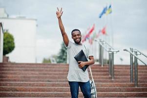 estudante africano masculino posou com mochila e itens escolares no pátio da universidade, contra bandeiras de diferentes países. mostrar dois dedos. foto