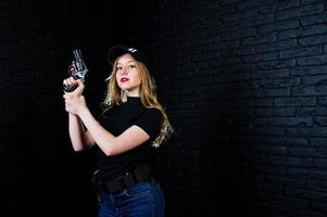 agente feminina do FBI no boné e com arma no estúdio contra a parede de tijolos escuros. foto