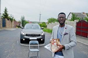 homem afro-americano rico e elegante de blazer e calça branca, óculos lê revista contra seu carro suv. foto