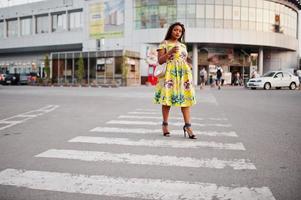 linda garota afro-americana de pequena altura com dreadlocks, use um vestido amarelo colorido, andando na faixa de pedestres contra o centro comercial com uma xícara de café à mão. foto