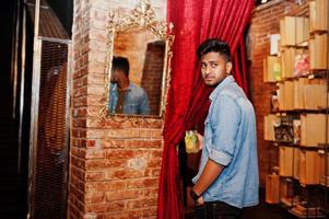 retrato de bonito bem sucedido barbudo sul-asiático, jovem indiano freelancer em camisa jeans azul em pé no café com coquetel contra espelho. foto