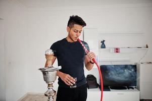 homem indiano bonito e elegante em pé de preto no quarto, fumando cachimbo de água com óculos na mão. foto