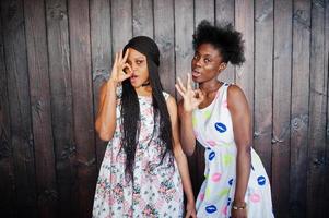 duas namoradas africanas negras em vestidos de verão posaram contra um fundo escuro de madeira e mostrando sinal de dedos bem. foto