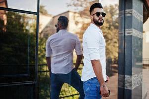 elegante modelo de homem árabe alto na camisa branca, jeans e óculos de sol posou na rua da cidade. barba cara árabe atraente contra edifício moderno. foto