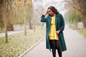 mulher afro-americana elegante no casaco verde e vestido amarelo posou contra o parque outono. foto