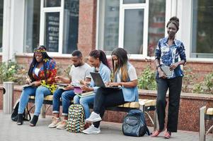 grupo de cinco estudantes universitários africanos passando tempo juntos no campus no pátio da universidade. amigos afro negros estudando no banco com itens escolares, notebooks laptops. foto