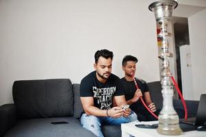 dois homens indianos bonitos e elegantes relaxando no quarto e fumando narguilé. um deles olhando para o celular. foto