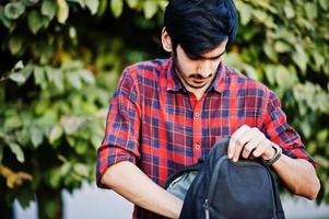 homem jovem estudante indiano na camisa xadrez vermelha e jeans com mochila posou na rua. foto