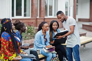 grupo de cinco estudantes universitários africanos passando tempo juntos no campus no pátio da universidade. amigos afro negros estudando no banco com itens escolares, notebooks laptops. foto