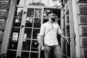 elegante modelo de homem árabe alto na camisa branca, jeans e óculos de sol posou na rua da cidade. barba cara árabe atraente contra a porta de entrada do restaurante. foto