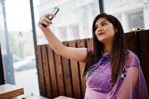 garota hindu indiana no tradicional saree violeta sentado à mesa de café com o celular nas mãos, fazendo selfie. foto