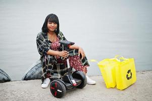 linda mulher afro-americana sente-se perto de segway ou hoverboard. garota negra com símbolo de reciclagem de sacos ecológicos de pano amarelo. foto