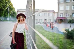 menina modelo loira elegante e bonita na saia de veludo vermelho elegante, blusa branca e chapéu, posou com telefone e fones de ouvido. foto