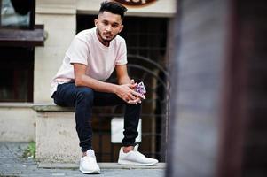 homem de barba indiana elegante na camiseta rosa. modelo da índia sentado com o celular nas mãos ao ar livre nas ruas da cidade. foto