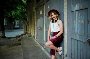 menina modelo loira elegante e bonita na saia de veludo vermelho elegante, blusa branca e chapéu, posou com telefone e fones de ouvido. foto