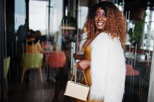 glamour mulher afro-americana de vestido amarelo e capa de lã branca com bolsa posada na entrada da porta de vidro do restaurante. foto