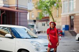 linda e magra garota afro-americana de vestido vermelho com dreadlocks posou ao ar livre no carro branco pequeno de fundo de rua. elegante modelo preto. foto