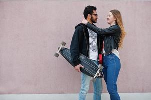 legal casal multirracial posando contra a parede rosa com longboard. foto