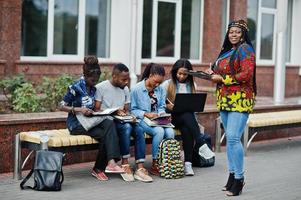 grupo de cinco estudantes universitários africanos passando tempo juntos no campus no pátio da universidade. amigos afro negros estudando no banco com itens escolares, notebooks laptops. foto