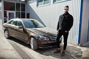 moda homem árabe usar jaqueta jeans preta e óculos escuros posou contra carro moderno de negócios. cara modelo árabe elegante, bem sucedido e elegante. foto