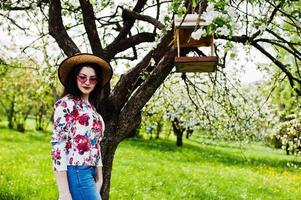 retrato de primavera de menina morena de óculos cor de rosa e chapéu no jardim de flor verde. foto