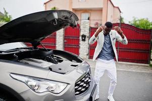 homem afro-americano elegante e rico fica na frente de um carro suv quebrado precisa de assistência olhando sob o capô aberto. foto