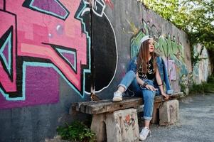 menina elegante casual hipster em boné e jeans usa música de fones de ouvido do telefone celular contra grande parede de graffiti com bomba. foto