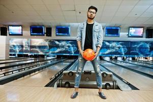 homem asiático de barba elegante na jaqueta jeans e óculos em pé na pista de boliche com bola na mão. foto