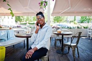 garota afro-americana usa óculos com celular sentado no caffe ao ar livre. foto