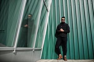 moda barba rica homem árabe usar jaqueta jeans preta e óculos de sol posou contra a parede verde com janelas. cara modelo árabe elegante, bem sucedido e elegante. foto