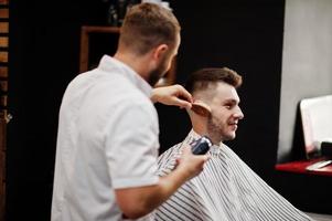 jovem barbudo cortando o cabelo pelo cabeleireiro enquanto está sentado na cadeira na barbearia. alma de barbeiro. foto