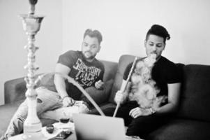 dois homens indianos bonitos e elegantes relaxando no quarto e fumando narguilé. foto