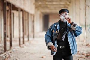 homem afro-americano na jaqueta jeans, boina e óculos, fumando charuto na fábrica abandonada. foto