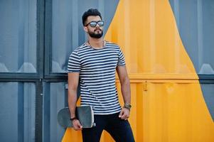 modelo de homem de barba árabe alto bonito na camisa despojada posou ao ar livre. cara árabe na moda em óculos de sol com skate contra parede pintada de amarelo. foto