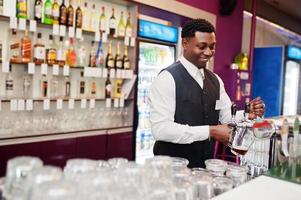 barman americano africano no bar derramando da torneira de cerveja fresca no copo no pub. foto