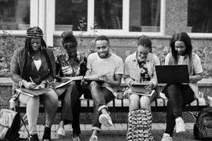 grupo de cinco estudantes universitários africanos passando tempo juntos no campus no pátio da universidade. amigos afro negros estudando no banco com itens escolares, notebooks laptops. foto