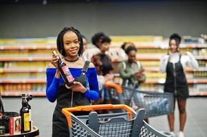mulher africana escolhe vinho no supermercado contra seus amigos afro com carrinho de compras. foto