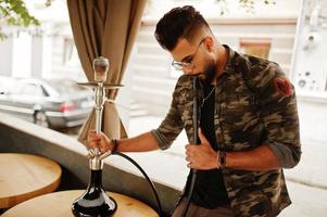 homem árabe de barba elegante de óculos e jaqueta militar fumando cachimbo de água no bar da rua. modelo árabe descansando. foto