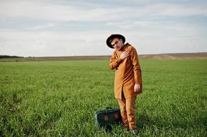 homem elegante de óculos, jaqueta marrom e chapéu com bolsa posou em campo verde. foto