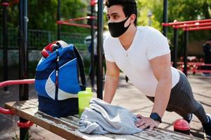 retrato esportivo homem árabe com máscara facial médica preta fazendo exercícios de treino no local da academia ao ar livre durante a quarentena de coronavírus. foto