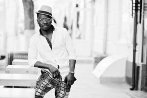 homem afro-americano elegante na camisa branca e calça colorida com chapéu e óculos posou ao ar livre. menino modelo preto elegante. foto