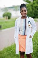 jovem médica afro-americana de jaleco branco com um estetoscópio posou ao ar livre. foto