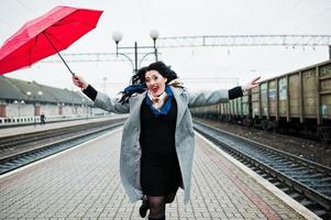 menina morena de casaco cinza com guarda-chuva vermelho na estação ferroviária. foto
