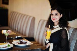 jovem indiana usar no elegante saree preto sentado no restaurante com um copo de suco na mão. foto