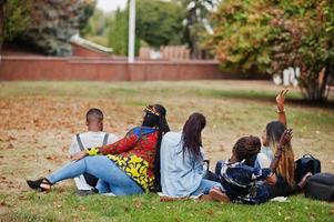 parte de trás do grupo cinco estudantes universitários africanos passando tempo juntos no campus no pátio da universidade. amigos negros afro sentados na grama e estudando com laptops. foto