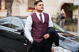 elegante empresário indiano em traje formal colete em pé contra carro preto na rua da cidade. foto